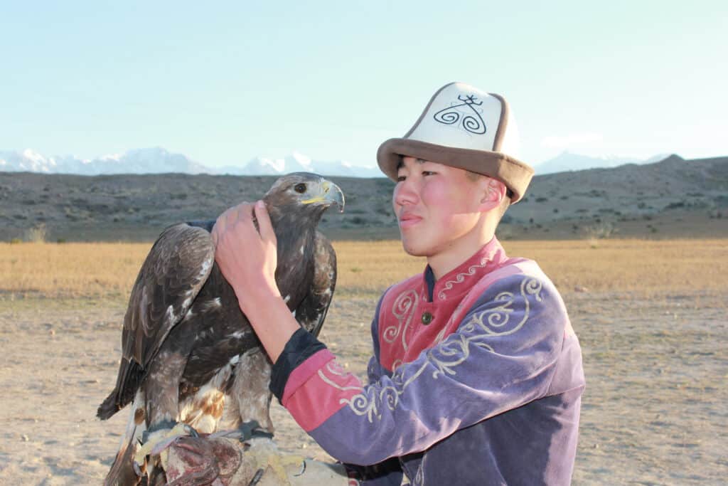 Junger Adlerjäger in traditioneller kirgisischer Kleidung mit Steinadler vor Bergkulisse