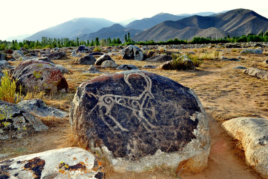 Fels mit Tier-Petroglyphe in Cholpon Ata, dahinter weite Ebene und Berge im Abendlicht